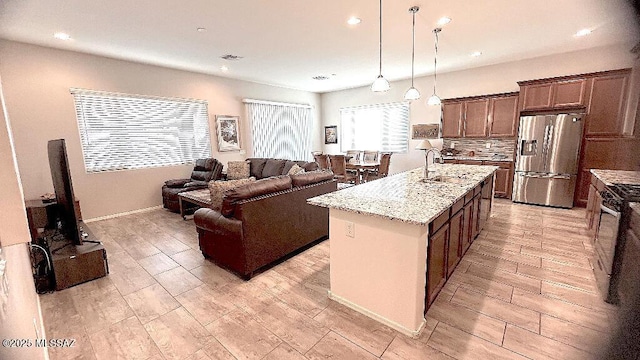 kitchen with light stone countertops, stainless steel fridge, tasteful backsplash, a kitchen island with sink, and decorative light fixtures