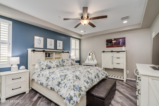 bedroom featuring dark wood-type flooring, a tray ceiling, ceiling fan, and a barn door