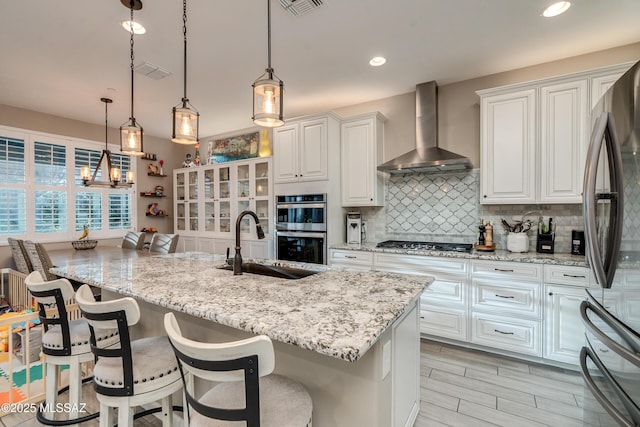 kitchen featuring pendant lighting, decorative backsplash, white cabinetry, an island with sink, and wall chimney exhaust hood