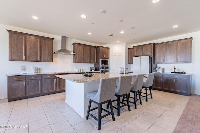 kitchen featuring a breakfast bar area, stainless steel microwave, freestanding refrigerator, dark brown cabinetry, and wall chimney exhaust hood