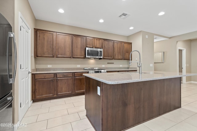 kitchen with light tile patterned floors, sink, stainless steel appliances, light stone countertops, and a center island with sink