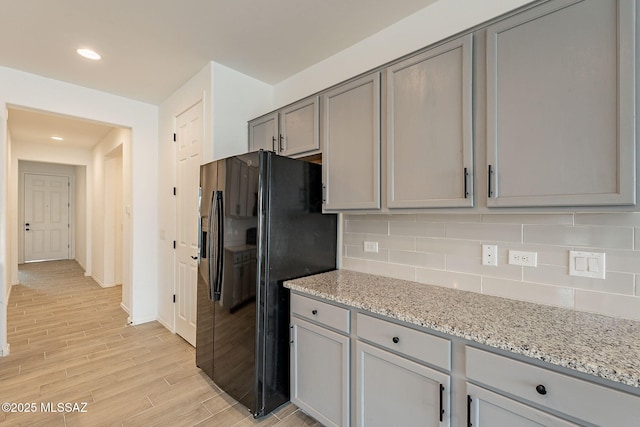 kitchen with gray cabinetry, light stone countertops, black fridge, decorative backsplash, and light wood-type flooring