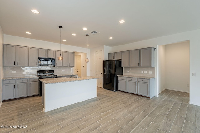 kitchen featuring pendant lighting, black appliances, a center island with sink, tasteful backsplash, and light stone counters