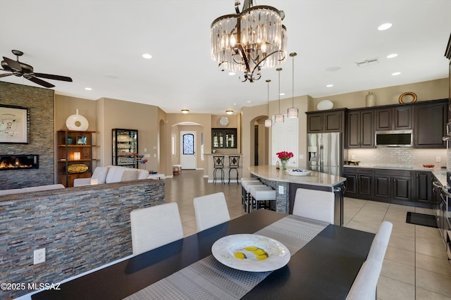 tiled dining room with ceiling fan with notable chandelier and a stone fireplace