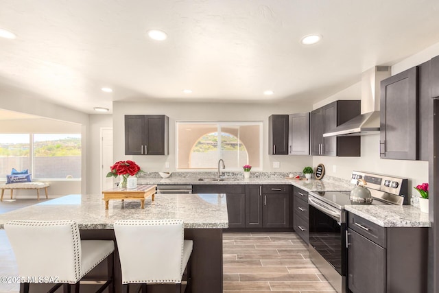 kitchen featuring stainless steel electric stove, a breakfast bar, sink, light stone counters, and wall chimney range hood