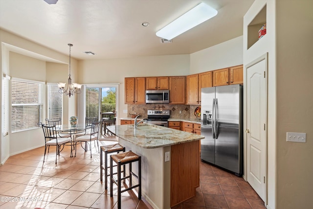 kitchen with stainless steel appliances, tasteful backsplash, light stone counters, decorative light fixtures, and a center island with sink