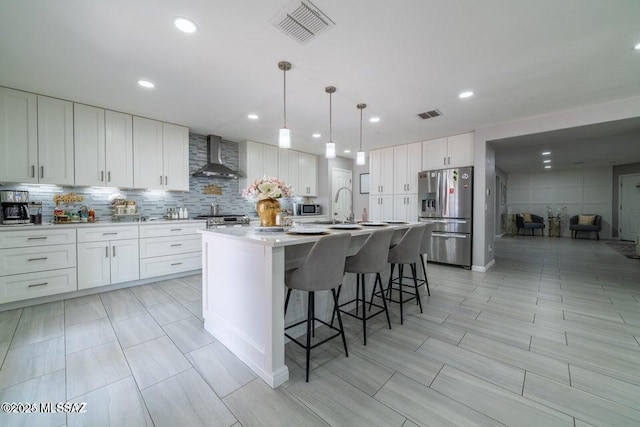 kitchen with wall chimney range hood, a center island with sink, white cabinetry, hanging light fixtures, and appliances with stainless steel finishes