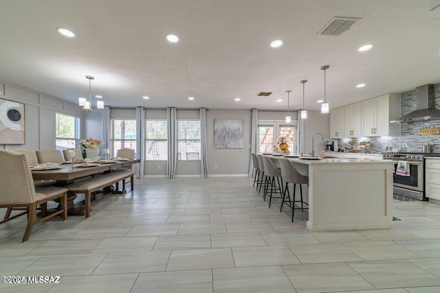 kitchen featuring hanging light fixtures, an island with sink, stainless steel range oven, and wall chimney range hood