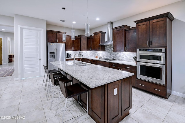 kitchen featuring sink, a kitchen island with sink, light stone counters, stainless steel appliances, and wall chimney range hood