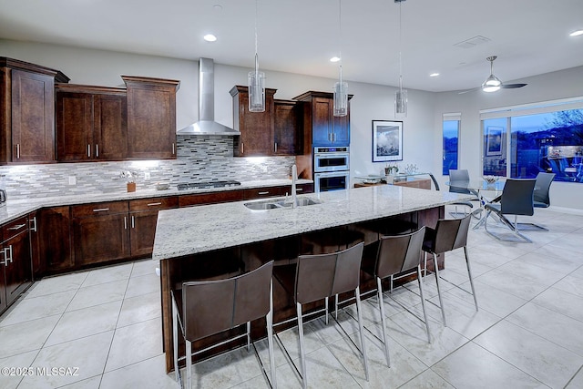 kitchen featuring an island with sink, light stone countertops, sink, and wall chimney exhaust hood