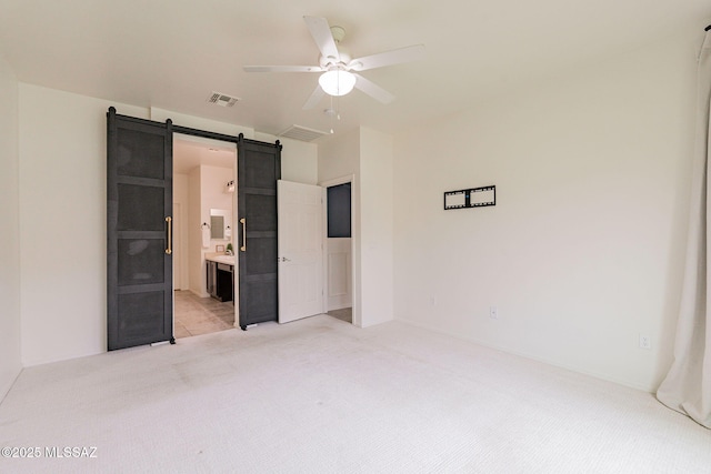 unfurnished bedroom featuring ceiling fan, a barn door, light colored carpet, and connected bathroom