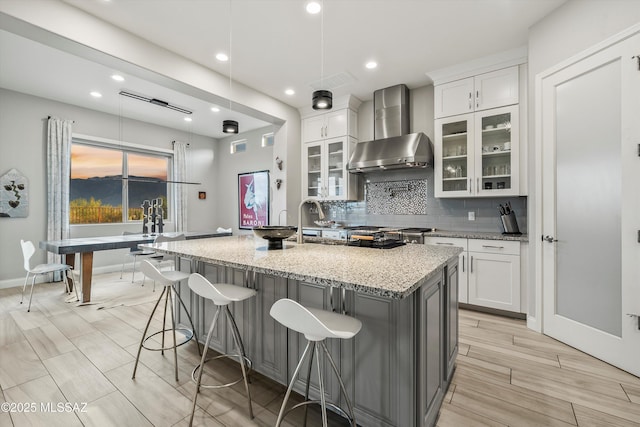 kitchen featuring white cabinets, light stone countertops, wall chimney exhaust hood, and an island with sink