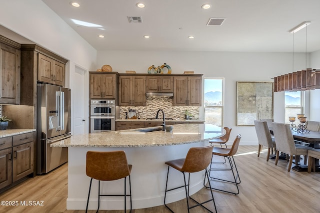 kitchen with light hardwood / wood-style floors, a center island with sink, appliances with stainless steel finishes, light stone counters, and sink