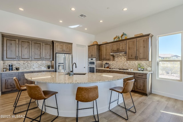 kitchen with light hardwood / wood-style floors, sink, a kitchen island with sink, appliances with stainless steel finishes, and light stone counters