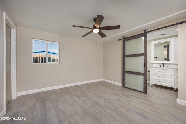 unfurnished bedroom with ensuite bath, ceiling fan, sink, a barn door, and light hardwood / wood-style floors