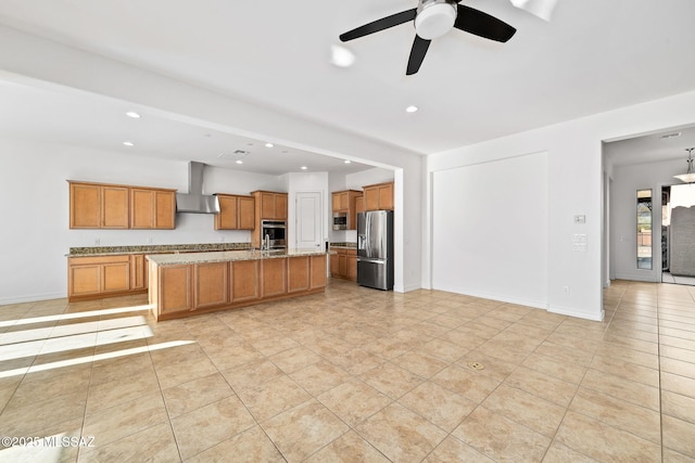 kitchen with a center island with sink, wall chimney exhaust hood, ceiling fan, light stone countertops, and appliances with stainless steel finishes