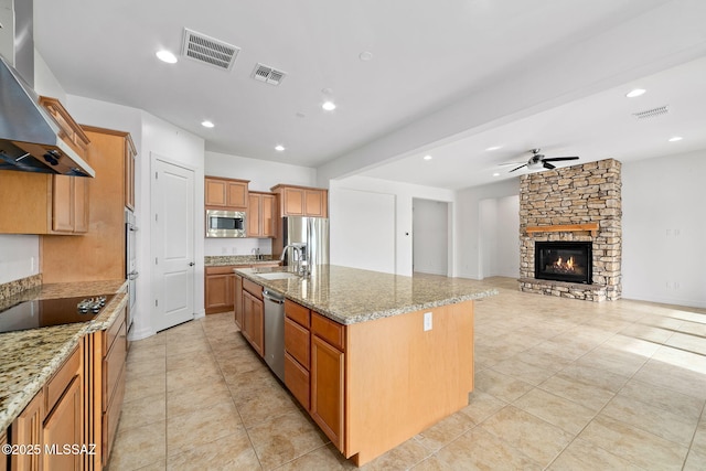 kitchen featuring a kitchen island with sink, wall chimney exhaust hood, ceiling fan, appliances with stainless steel finishes, and light stone counters