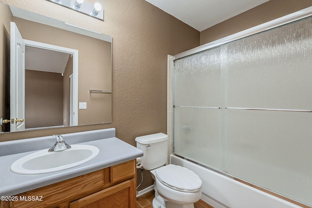 full bathroom featuring combined bath / shower with glass door, a textured wall, vanity, and toilet