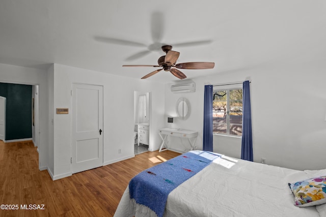 bedroom with a ceiling fan, a wall mounted AC, ensuite bath, wood finished floors, and baseboards