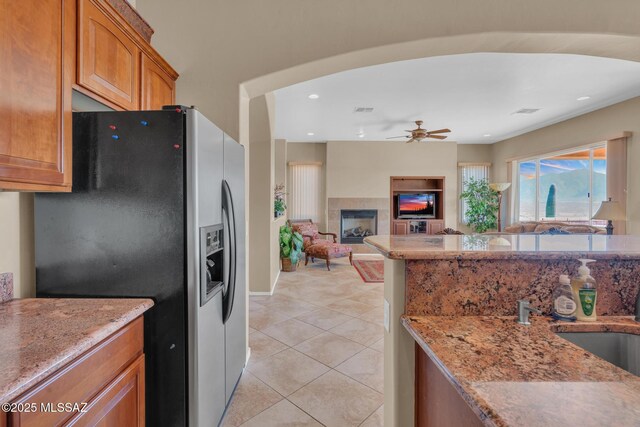 kitchen with ceiling fan, a fireplace, light tile patterned flooring, stainless steel fridge with ice dispenser, and light stone counters