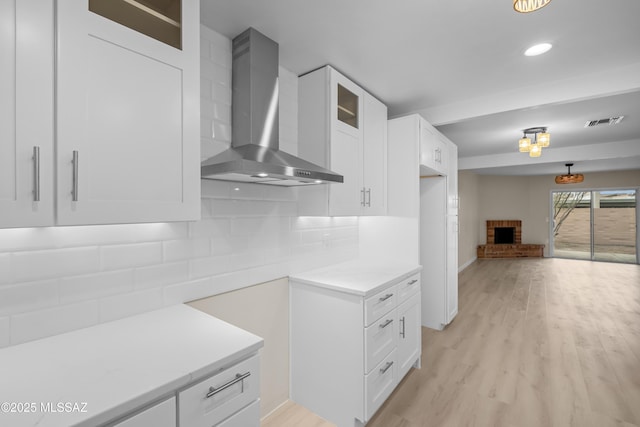 kitchen with white cabinetry, a fireplace, light hardwood / wood-style floors, decorative backsplash, and wall chimney range hood