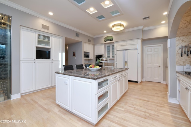 kitchen featuring appliances with stainless steel finishes, light wood-type flooring, dark stone counters, white cabinetry, and an island with sink