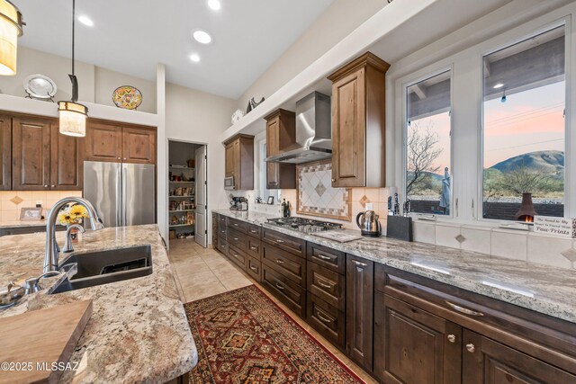 kitchen featuring hanging light fixtures, wall chimney exhaust hood, light stone countertops, appliances with stainless steel finishes, and dark brown cabinets