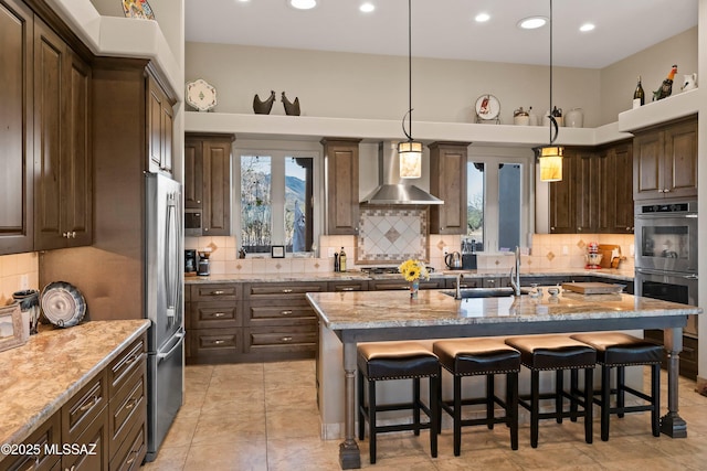 kitchen featuring a kitchen island with sink, stainless steel appliances, wall chimney range hood, decorative backsplash, and a kitchen bar