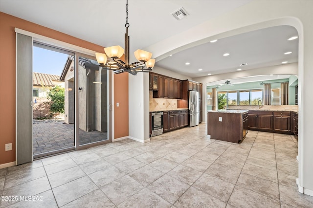 kitchen featuring ceiling fan with notable chandelier, decorative backsplash, dark brown cabinetry, a kitchen island, and stainless steel fridge