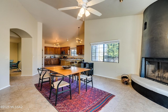 dining area with light tile patterned flooring, a large fireplace, ceiling fan with notable chandelier, and high vaulted ceiling