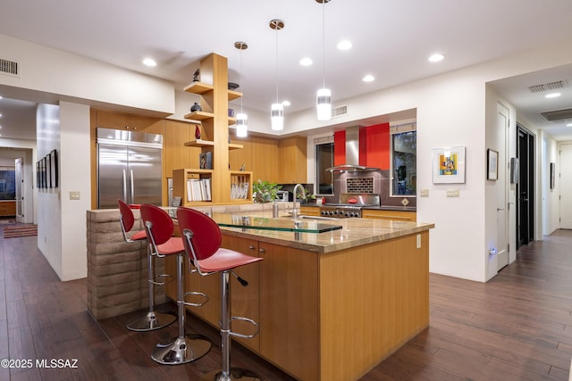 kitchen with light stone countertops, stainless steel built in refrigerator, wall chimney range hood, pendant lighting, and range