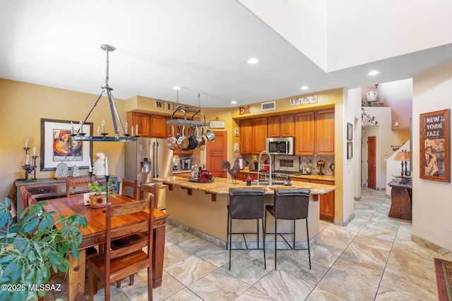 kitchen with a center island with sink, hanging light fixtures, a breakfast bar area, light stone countertops, and stainless steel appliances