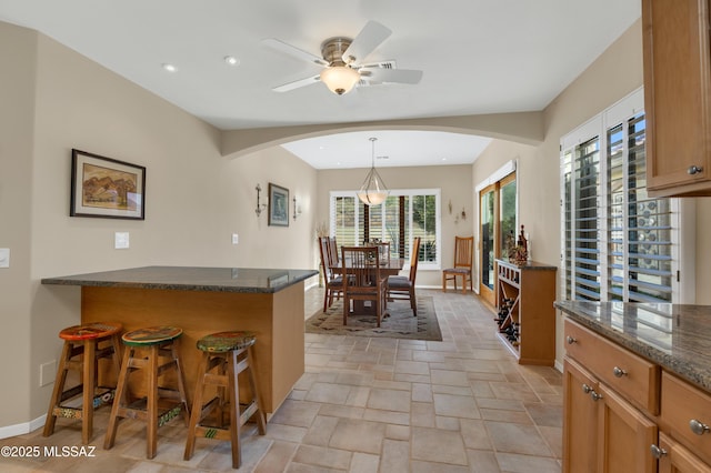 kitchen featuring ceiling fan, a kitchen bar, hanging light fixtures, and dark stone countertops