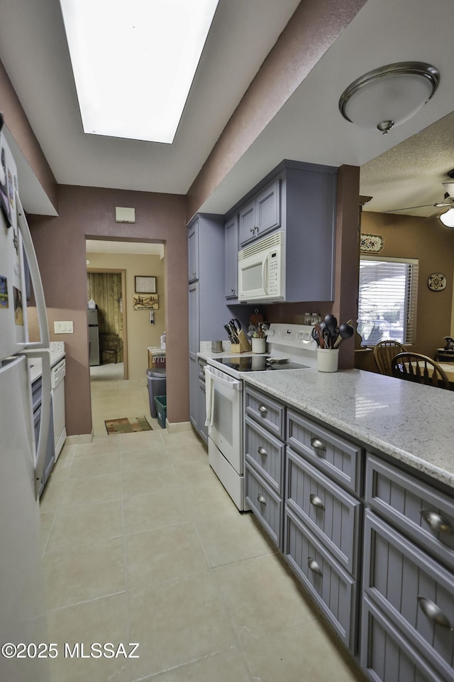 kitchen with gray cabinetry, light stone countertops, ceiling fan, a skylight, and white appliances