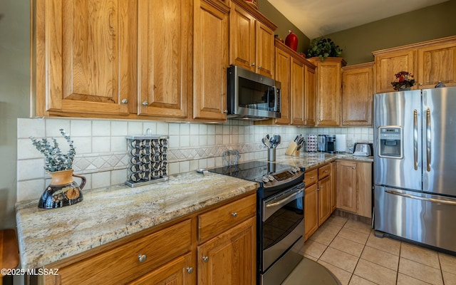 kitchen featuring light tile patterned floors, decorative backsplash, brown cabinetry, appliances with stainless steel finishes, and light stone counters