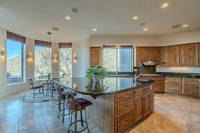 kitchen featuring decorative light fixtures, stainless steel gas cooktop, dark stone countertops, tasteful backsplash, and an island with sink