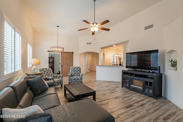 living room featuring a barn door, a towering ceiling, ceiling fan with notable chandelier, and light hardwood / wood-style floors