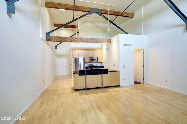 kitchen featuring light brown cabinetry, light wood-type flooring, appliances with stainless steel finishes, beam ceiling, and a high ceiling