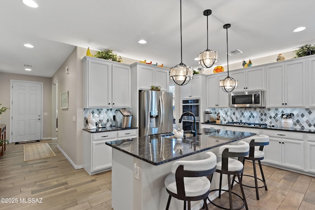 kitchen with stainless steel appliances, white cabinets, decorative backsplash, hanging light fixtures, and a center island with sink