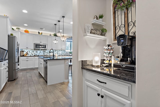 kitchen featuring dark stone countertops, stainless steel appliances, an island with sink, white cabinetry, and decorative light fixtures