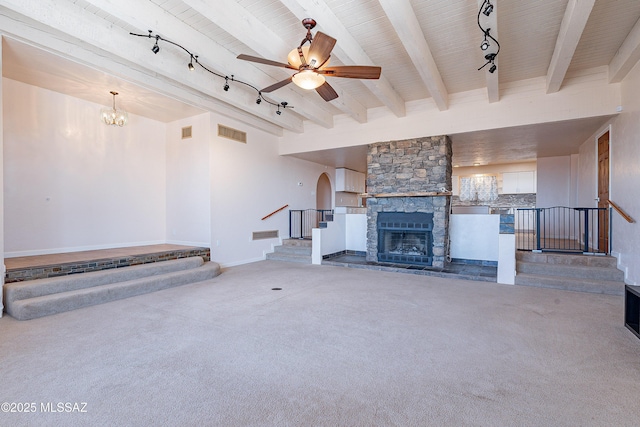 unfurnished living room featuring rail lighting, ceiling fan with notable chandelier, beamed ceiling, carpet floors, and a stone fireplace