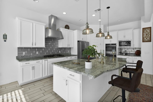 kitchen featuring wall chimney exhaust hood, tasteful backsplash, decorative light fixtures, a kitchen island with sink, and appliances with stainless steel finishes