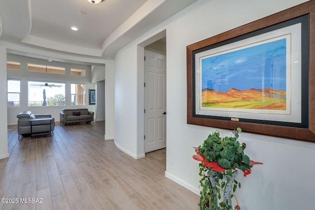 hallway featuring light wood-style floors, a tray ceiling, baseboards, and recessed lighting