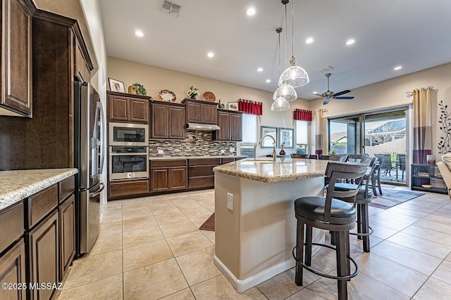 kitchen with stainless steel appliances, an island with sink, decorative backsplash, light stone counters, and a breakfast bar