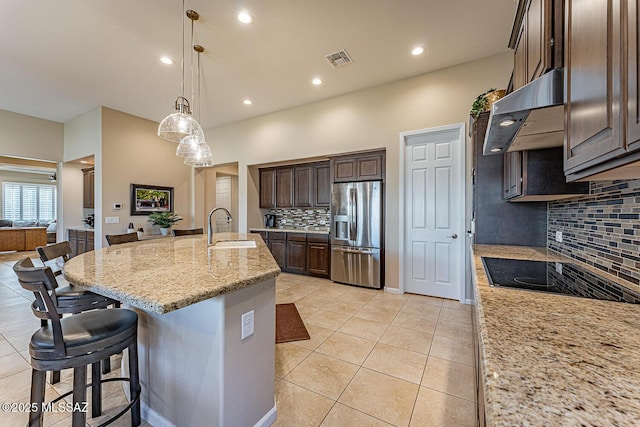 kitchen featuring sink, range hood, stainless steel fridge with ice dispenser, an island with sink, and light stone countertops