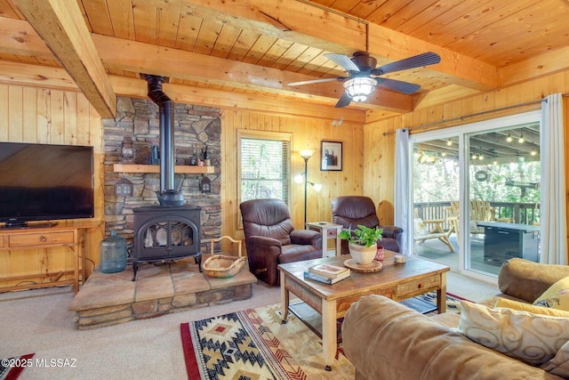 carpeted living room featuring beam ceiling, ceiling fan, wooden walls, and a wood stove