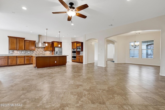 kitchen with stainless steel appliances, an island with sink, hanging light fixtures, and wall chimney range hood