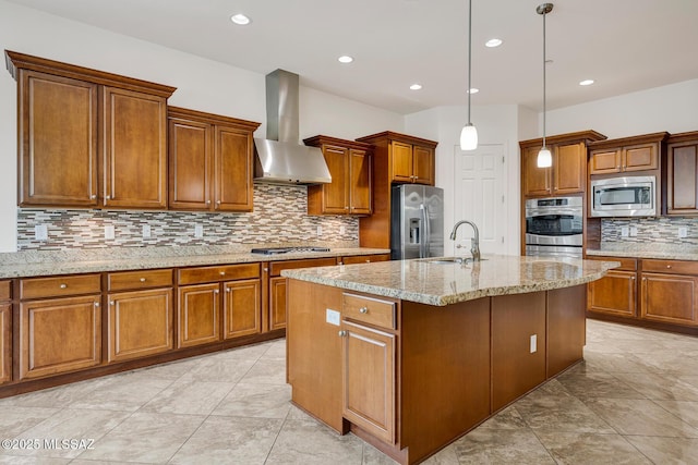 kitchen featuring wall chimney range hood, sink, appliances with stainless steel finishes, an island with sink, and decorative light fixtures