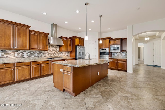 kitchen featuring wall chimney range hood, appliances with stainless steel finishes, a kitchen island with sink, hanging light fixtures, and light stone counters