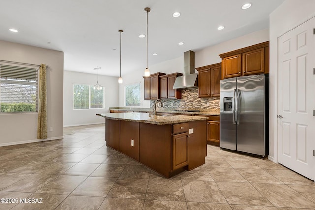 kitchen with appliances with stainless steel finishes, an island with sink, pendant lighting, light stone countertops, and wall chimney range hood